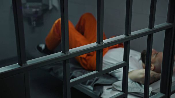 Inmate in orange uniform lies on prison cell bed. Prisoner serves imprisonment term for crime. Criminal in detention center, correctional facility. Justice system. View through metal bars. Dolly shot. - Photo, Image