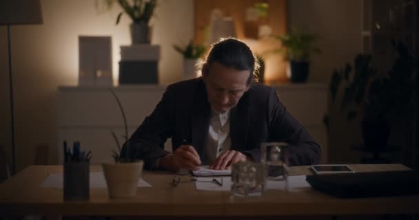 Ανώτερος διευθυντής άντρας γράφει στο ημερολόγιο στο γραφείο ενώ εργάζεται αργά το βράδυ στο γραφείο - Πλάνα, βίντεο