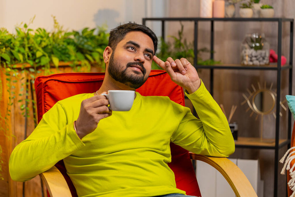 Atraktivní usměvavý Ind pije šálek teplé kávy nebo bylinkový čaj sedí doma v obývacím pokoji houpací křeslo v dopoledních hodinách. Arabský chlap si užívá relaxaci sám, odpočívá, dává si pauzu - Fotografie, Obrázek