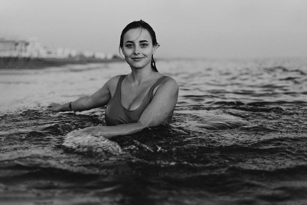 Een jonge vrouw met een glimlach op haar gezicht zwemt 's avonds vroeg in het rustige oceaanwater. Haar hoofd en schouders zijn zichtbaar boven de waterlijn - Foto, afbeelding