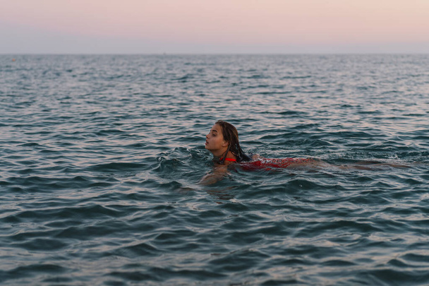 Mladá žena s úsměvem na tváři se během večera koupe v klidných vodách oceánu. Nad hladinou je vidět její hlava a ramena. - Fotografie, Obrázek
