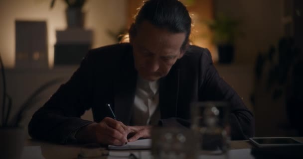 Uomo d'affari anziano che scrive nel diario alla scrivania mentre lavora fino a tarda notte sul posto di lavoro - Filmati, video