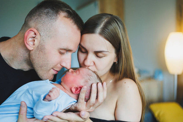 Szczęśliwi młodzi rodzice trzymający swoje noworodki w ramionach. Romper na nowonarodzonego chłopca. Noworodek w rękach mamy i taty - Zdjęcie, obraz