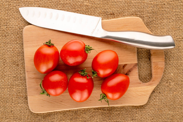 Kilka pomidorów koktajlowych z nożem i drewnianą deską kuchenną na ściereczce z juty, makro, widok z góry. - Zdjęcie, obraz