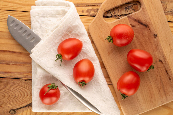 Several cocktail tomatoes with a knife on a white napkin and a wooden kitchen board on a wooden table, macro, top view. - Photo, Image