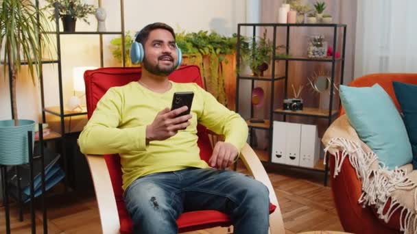 Evde koltukta dans eden kablosuz kulaklıklı mutlu, gevşemiş Hint Arap adam akıllı telefon eğlencesinde favori, enerjik disko pop müziğini seçiyor. İnsanlar hafta sonu aktiviteleri - Video, Çekim