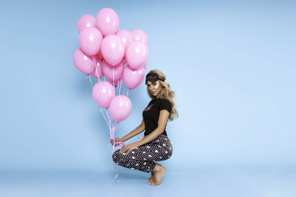 Piękna młoda, szczęśliwa dziewczyna w seksownej satynowej piżamie trzyma różowe balony i zmysłowo pozuje na niebieskim pastelowym tle. moda piżama. - Zdjęcie, obraz
