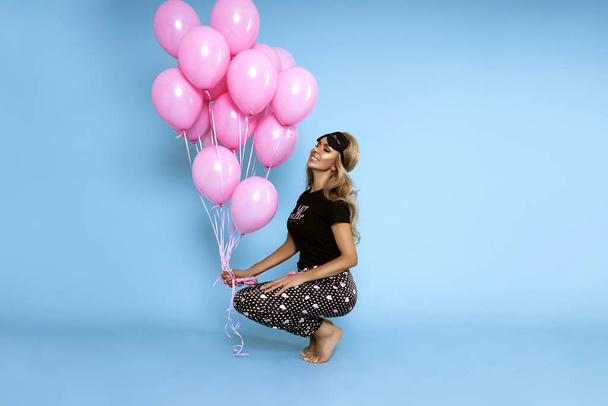 Beautiful young happy girl in sexy satin pajamas holds pink balloons and poses sensually on a blue pastel background. Pajama fashion. - Photo, Image