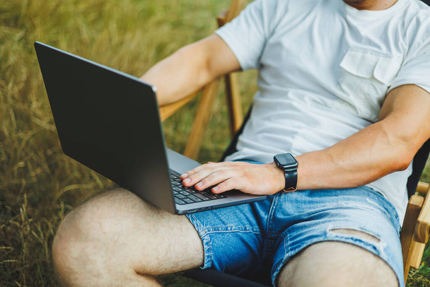 Un uomo su una sedia pieghevole in legno si siede in natura e lavora in remoto online su un computer portatile. Il concetto di svago nella sola natura. - Foto, immagini