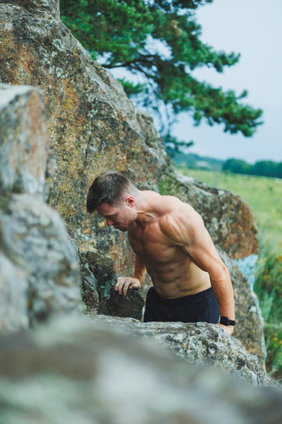 Lihaksikas nuori mies istuu kallioilla lähellä metsää. Miehen helpotus ja lihaksikas ruumiinrakenne. - Valokuva, kuva