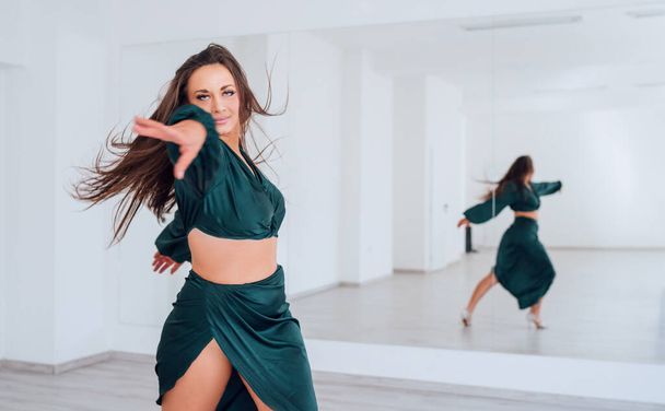 Graceful woman dressed in dark green Latin dancing dress doing elegant dance pas in white color big hall with big mirror wall. People's expressions during dancing, beauty of woman's body concept image - Фото, изображение