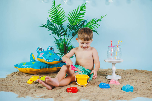 Zona foto estiva per bambini. Un bambino in pantaloncini si siede sulla sabbia e gioca con i giocattoli. Vacanze estive. - Foto, immagini