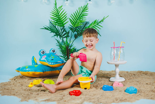 Summer photo zone for children. A little boy in shorts sits on the sand and plays with toys. Summer vacation. - Photo, Image