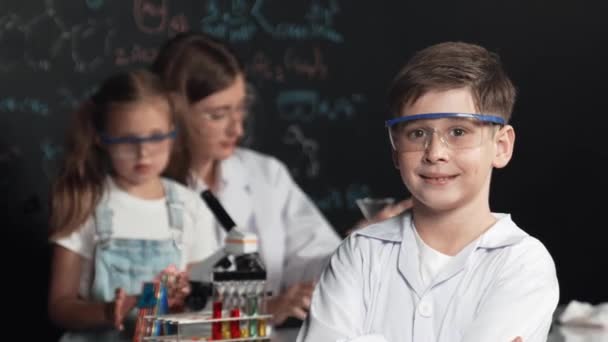 Öğretmen ve arkadaşı STEM fen dersinde deney yaparken akıllı çocuk kollarını kavuşturuyor. Öğrenci renkli çözeltileri karıştırırken laboratuvarda tahtada duran yazılı kimya teorisi. Etkinlik. - Video, Çekim