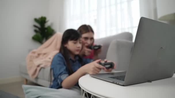 Onnellinen tyttö pelaa peliä ja istuu, kun äiti makaa sohvalla kannettavan tietokoneen näytöllä. Valkoihoinen vanhempi ja houkutteleva tyttö viettää aikaa yhdessä pitäen joystick ja keskittyä voittaa pelejä. Pedagogiikka. - Materiaali, video