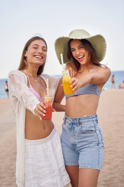 Vertical portrait of two caucasian girls friends looking at the camera and toasting with smoothies on the beach having fun and smiling happy enjoying the summer on their vacation at the sea together - Photo, Image