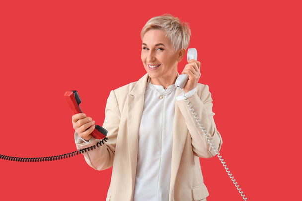 volwassen vrouw met twee telefoon ontvangers op rode achtergrond - Foto, afbeelding