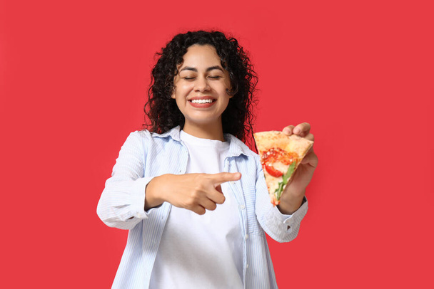 Belle jeune femme afro-américaine heureuse pointant du doigt une tranche de délicieuse pizza sur fond rouge - Photo, image