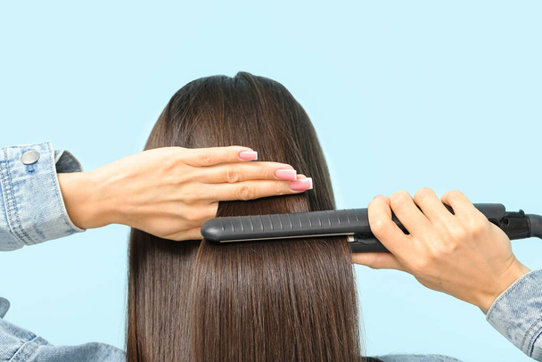 Beautiful young woman straightening hair on blue background, back view - Photo, Image