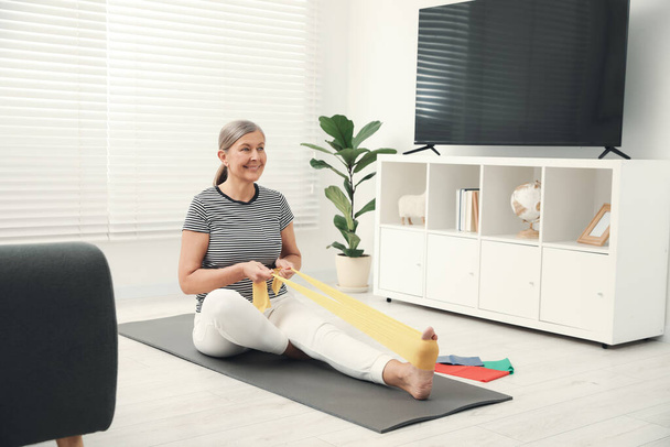 Senior woman doing exercise with fitness elastic band on mat at home - Valokuva, kuva