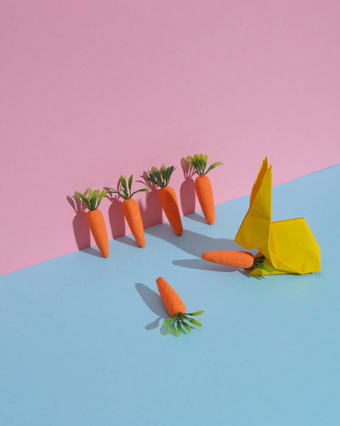 Conejo en origami con zanahorias sobre fondo azul-rosado. Diseño creativo de Pascua o primavera - Foto, imagen