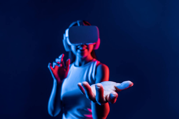Inteligentna kobieta stojąca otoczona światłem neonowym nosi zestaw słuchawkowy VR łączący metawertyczną, futurystyczną technologię społeczności cyberprzestrzeni. Elegancka kobieta używa ręcznego trzymania wygenerowanego obiektu wirtualnego. omamy. - Zdjęcie, obraz