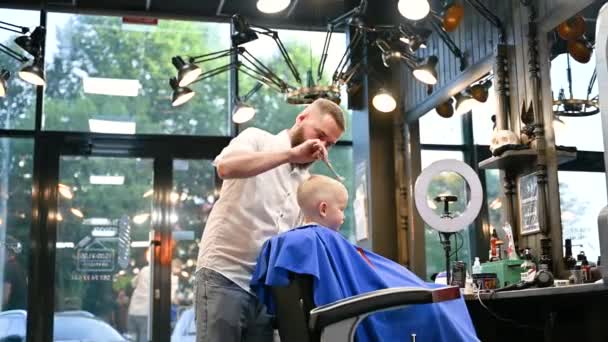 Peluquero hombre cortar el pelo de los niños pequeños usando peine y tijeras. A un niño le cortan el pelo hombres adultos, probablemente peluquero. Peluquería profesional y cliente lindo en la barbería moderna. - Imágenes, Vídeo