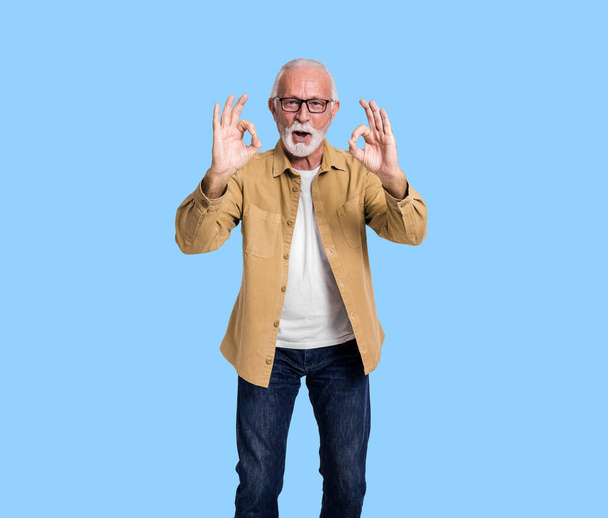 Ritratto di imprenditore anziano di sesso maschile che mostra segni OK e urla allegramente su sfondo blu - Foto, immagini