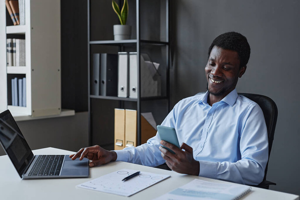 Πορτρέτο του χαμογελαστού μαύρου επιχειρηματία χρησιμοποιώντας smartphone κάθεται στο γραφείο στο γραφείο, αντίγραφο χώρου - Φωτογραφία, εικόνα