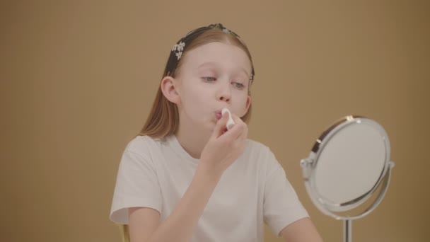 Pretty Caucasian teenage girl applying lip balm. Beautiful blonde girl looks in the mirror and makes lip care at home. The daily chore of cracking. Influencer Beauty Ad Concept. Beige backdrop  - Footage, Video