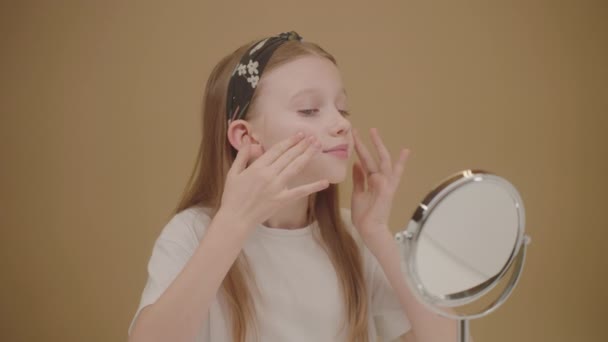 素敵な若い女の子はフェイスクリームを着ています. かわいいブロンドの女の子は鏡を見て,頬に保湿剤を加えます. 輝く肌で純粋に。 十代の若者とインフルエンサーのための美しさとスキンケアの概念. 化粧品 広告. - 映像、動画