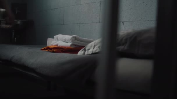 Prisonnier met uniforme orange et accessoires de salle de bains sur le lit en cellule de prison. Un criminel purge une peine d'emprisonnement en prison. Centre de détention ou établissement correctionnel. Vue à travers des barres métalliques. - Séquence, vidéo