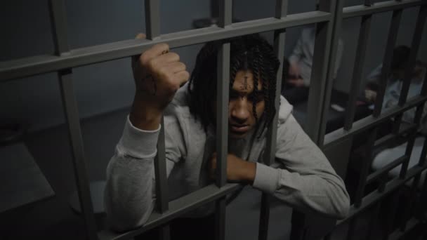 Boze Afro-Amerikaanse tiener gevangene staat in de gevangenis, houdt metalen tralies. Jonge gevangenen kaarten op bed op de achtergrond. Jeugddetentiecentrum of gevangenis. Hoge hoek. - Video