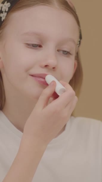 Zoom out effect. Funny blonde girl playing and having fun at home doing a beauty routine. Cheerful Caucasian teenage girl applies lip balm. Daily chapping routine. Beauty ads concept. Beige backdrop.  - Footage, Video