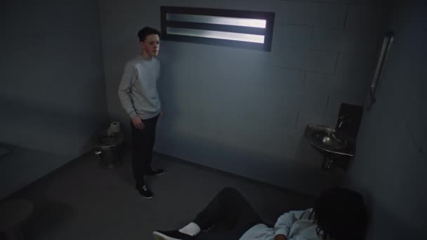 Diverse tieners praten terwijl ze in de gevangenis zitten. Jonge blanke gevangene staat bij het raam in de gevangenis cel, Afro-Amerikaanse gevangene zit op bed. Jeugdgevangenis. Bewakingscamera. - Video