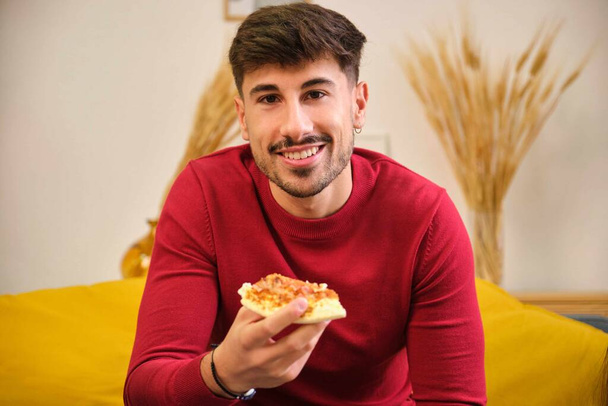 Kaukasische jongeman die pizza eet, glimlacht en naar de camera kijkt in een gedeelde flat. - Foto, afbeelding