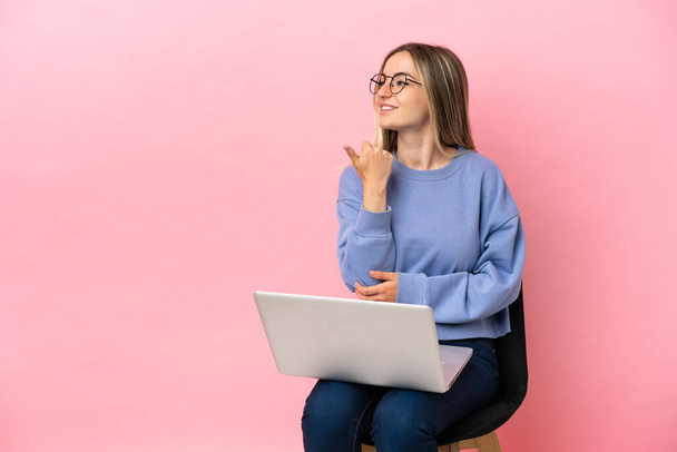 Jonge vrouw zit op een stoel met laptop over geïsoleerde roze achtergrond wijzend naar de zijkant om een product te presenteren - Foto, afbeelding