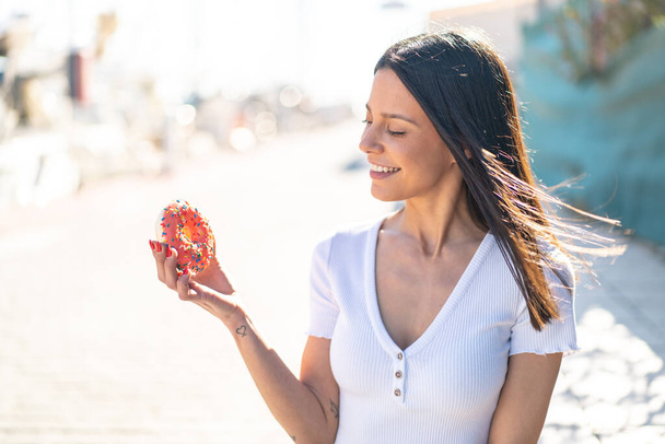 Νεαρή γυναίκα σε εξωτερικούς χώρους κρατώντας ένα ντόνατ σε εξωτερικούς χώρους με χαρούμενη έκφραση - Φωτογραφία, εικόνα