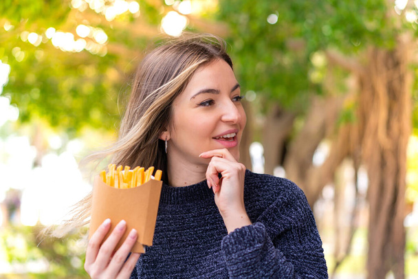 Young pretty Romanian woman holding fried chips at outdoors thinking an idea and looking side - Photo, Image