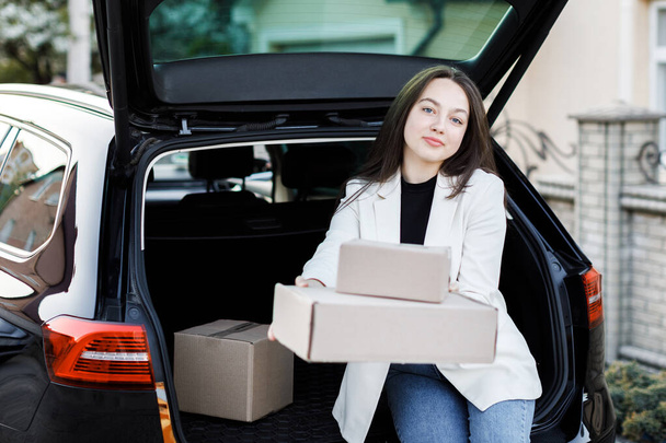 The girl sits in the trunk of the car and shows the boxes forward in front of the camera - Foto, afbeelding
