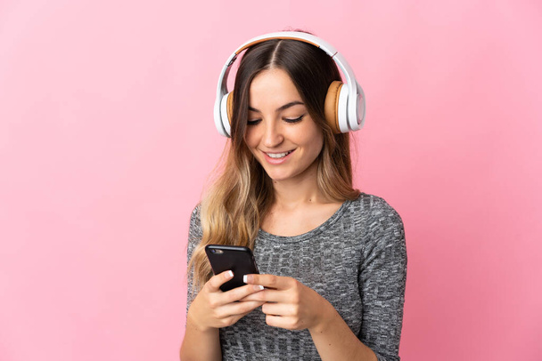 Junge Rumänin isoliert auf rosa Hintergrund Musik hören und auf Handy schauen - Foto, Bild