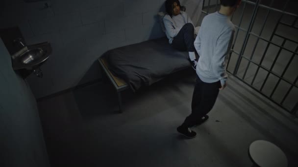 Kaksi monipuolista teini-ikäistä vankia puhuu vankilassa istumisesta. Yksi kävelee vankilan ympäri, toinen istuu sängyllä. Nuorisovankila tai vankila. CCTV-kamerakulma. - Materiaali, video