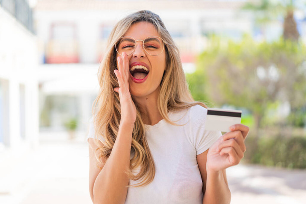 Bonita mujer uruguaya rubia sosteniendo una tarjeta de crédito al aire libre gritando con la boca abierta - Foto, Imagen