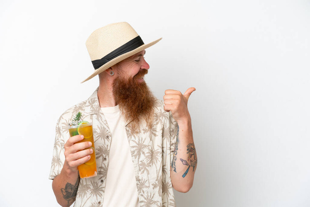 Kızıl saçlı, uzun sakallı, kumsalda kokteyl içen beyaz arka planda izole edilmiş bir adam bir ürünü sunmak için kenarı işaret ediyor. - Fotoğraf, Görsel