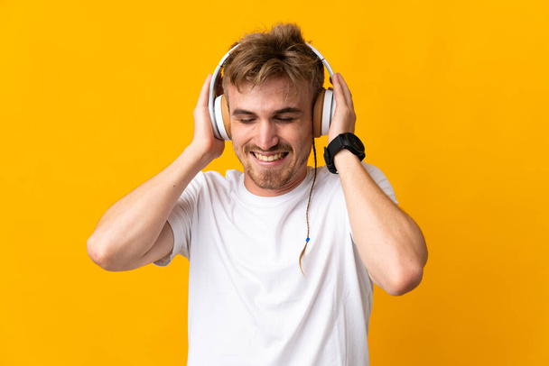 Jonge knappe blonde man geïsoleerd op gele achtergrond luisteren muziek - Foto, afbeelding