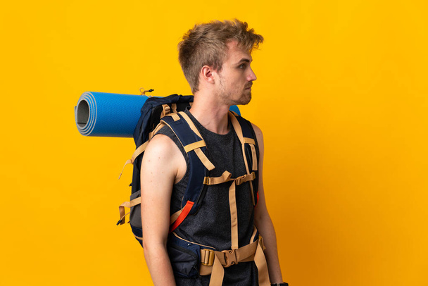 Mladý horolezec muž s velkým batohem izolované na žlutém pozadí při pohledu do strany - Fotografie, Obrázek