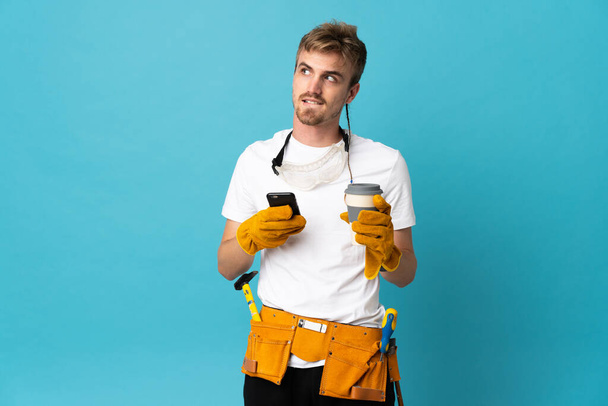 Jonge elektricien man over geïsoleerde muur met koffie om mee te nemen en een mobiele terwijl het denken iets - Foto, afbeelding