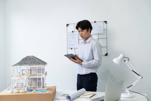 Profesjonalny przystojny azjatycki kierownik projektu za pomocą tabletu, aby sprawdzić model domu podczas kontroli struktury modelu domu starannie w biurze z planem powieszone za. Kreatywny projekt. Niepokalany. - Zdjęcie, obraz