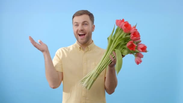 Ένας άνδρας στέκεται κρατώντας ένα ζωντανό μπουκέτο κόκκινες τουλίπες στα χέρια του, αναδεικνύοντας την ομορφιά των λουλουδιών σε ένα ουδέτερο σκηνικό - Πλάνα, βίντεο