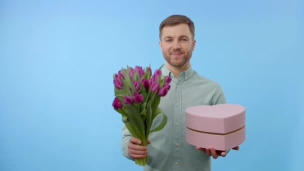 Muž držící kytici tulipánů a dárkové krabice, dívající se na kameru izolovanou na modrém pozadí. Koncept oslavy - Záběry, video